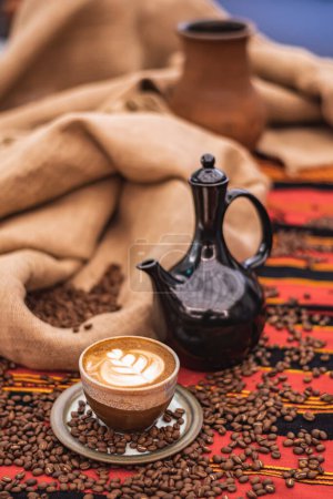 Téléchargez les photos : Cappuccino in a ceramic cup with flower or leaves drawn on the foam, teapot and roasted coffee beans or seeds scattered on a tablecloth with African decorations and in a jute sack, vertical - en image libre de droit