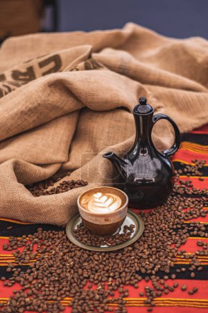 Téléchargez les photos : Cappuccino in a ceramic cup with flower or leaves drawn on the foam, teapot and roasted coffee beans or seeds scattered on a tablecloth with African decorations and in a jute sack, vertical - en image libre de droit
