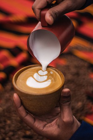 Téléchargez les photos : Black skin hand preparing a Cappuccino in a ceramic cup with flower or leaves drawn on the foam and roasted coffee beans or seeds scattered on a tablecloth with African decorations, vertical - en image libre de droit