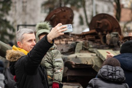 Téléchargez les photos : Kiev, Ukraine - 25 février 2023 : Père et fils prennent un selfie près d'une épave rouillée brûlée et fondue d'un char soviétique T-72B détruit lors de l'invasion russe de l'Ukraine à Kiev - en image libre de droit