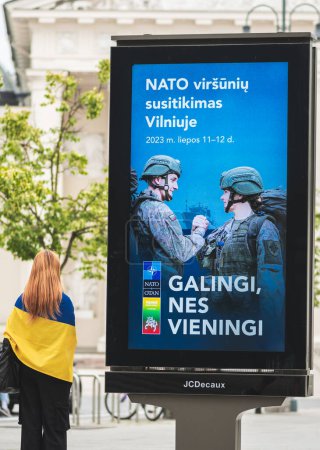 Téléchargez les photos : Vilnius, Lituanie - 22 juin 2023 : Bannière publicitaire informant sur le prochain sommet de l'OTAN 2023 dans le centre de Vilnius, capitale de la Lituanie avec une fille avec le drapeau de l'Ukraine marchant à proximité - en image libre de droit