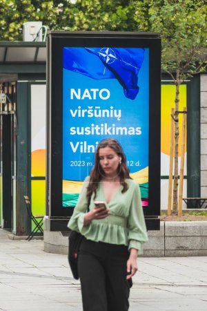 Photo for Vilnius, Lithuania - June 22 2023: Advertising banner informing about the upcoming Nato summit 2023 in the centre of Vilnius, capital of Lithuania with girl checking info walking nearby, vertical - Royalty Free Image