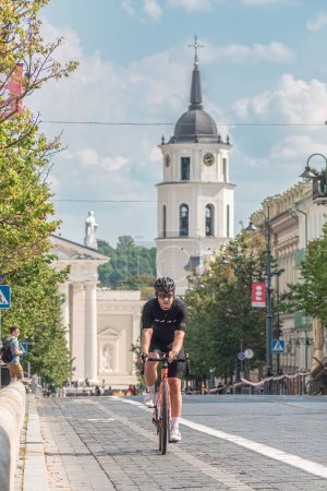 Photo for Vilnius, Lithuania - August 20 2023:  Boy riding a road bike with helmet on the bike road in the city centre of Vilnius, Lithuania with tower bell and cathedral on background, vertical - Royalty Free Image