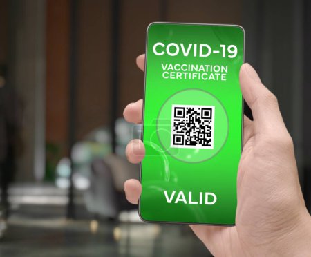 Photo for Corona Virus Tracking App concept with hand holding a cell phone with green "Valid Covid-19 Vaccination Certificate" mobile alert application design on screen with blurry Elegant Indoor background. - Royalty Free Image