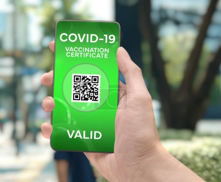 Foto de Corona Virus Tracking App concept with hand holding a cell phone with green "Valid Covid-19 Vaccination Certificate" mobile alert application design on screen in front of blurry urban park background. - Imagen libre de derechos