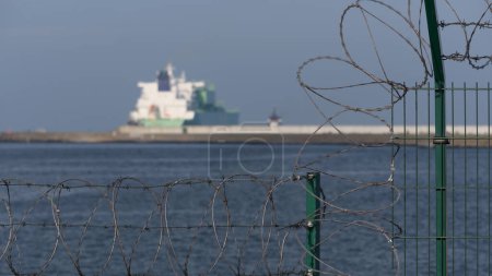 Téléchargez les photos : SITE DE PROTECTION - Clôture portuaire avec fil de rasoir et méthanier naviguant vers la mer - en image libre de droit