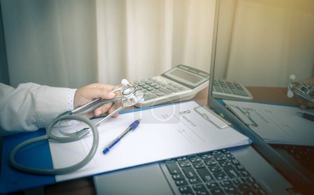 Photo for Doctor holding medical prescription paper clipboard with Stethoscope for patient note calcuator on Laptop computer, table in hospital Healthcare and medically concept, selective focus - Royalty Free Image