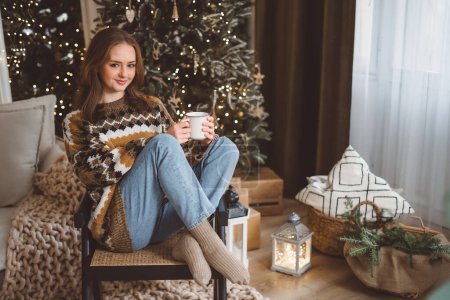 Téléchargez les photos : Gros plan portrait d'une jolie jeune femme portant un pull chaud avec une tasse rouge dans les mains sur le fond d'un arbre de Noël. La fille boit un verre dans un intérieur confortable du Nouvel An. - en image libre de droit