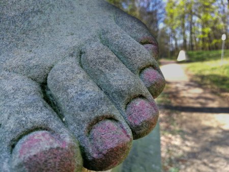 Téléchargez les photos : Pied nu d'une sculpture en grès avec des ongles rouges après une pédicure par les visiteurs du parc à la Weinbergschoelsschen à Dessau Grosskuehnau (Saxe-Anhalt). A côté d'elle un chemin dans le parc visible en arrière-plan. - en image libre de droit