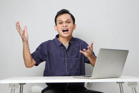 Foto de Enojado hombre asiático loco en el lugar de trabajo mientras está sentado frente a la computadora portátil. fondo aislado - Imagen libre de derechos
