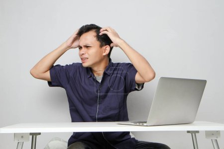 Foto de Confuso hombre asiático en el lugar de trabajo blanco sentado delante de la computadora portátil. fondo aislado - Imagen libre de derechos