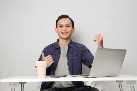 Foto de Alegre feliz asiático hombre en el lugar de trabajo sosteniendo una taza y dar pulgar hacia arriba signo mientras se sienta delante de la computadora portátil. sobre fondo aislado - Imagen libre de derechos