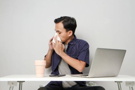 Foto de Asiático hombre tiene una secreción nasal, se limpia la nariz con papel tisú en el lugar de trabajo mientras está sentado frente a la computadora portátil. fondo aislado - Imagen libre de derechos
