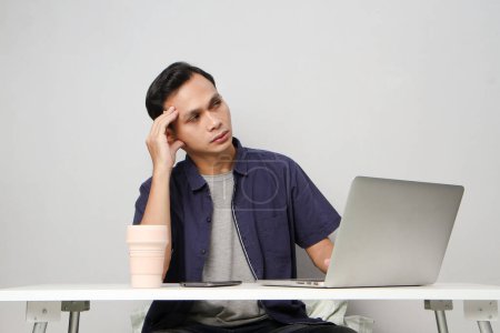 Foto de Pensativo confundido asiático hombre en el lugar de trabajo whiile sentado en frente de ordenador portátil. fondo aislado - Imagen libre de derechos