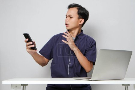 Foto de Hombre asiático enojado sosteniendo el teléfono móvil mientras está sentado frente a la computadora portátil. sobre fondos aislados - Imagen libre de derechos