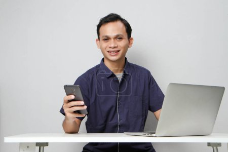 Foto de Feliz hombre asiático alegre sosteniendo el teléfono móvil mientras está sentado frente a la computadora portátil. sobre fondo aislado - Imagen libre de derechos