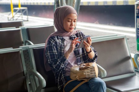 asiatique musulmane femme tenant smartphone assis sur banc à gare salle d'attente. concept de voyage