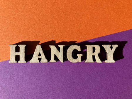 Téléchargez les photos : Hangry, un portmanteau ou mélangé mot mader de colère et faim. Lettres alphabet en bois isolées sur fond violet et orange - en image libre de droit