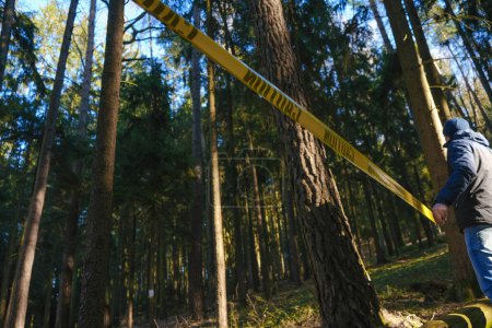 Photo for Crime scene. stop motion tape. Entry ban. Man pulling yellow barrier tape in pine dark forest. Murder and crime concept. - Royalty Free Image