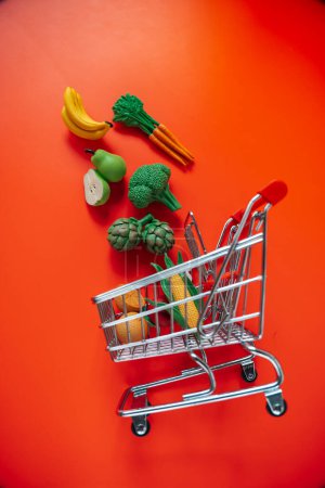 Lebensmittelverbraucherkorb.Lebensmittelkosten.Einkaufswagen mit Lebensmitteln auf rotem Hintergrund. Dekoratives Gemüse und Obst. Dekorativer Einkaufswagen mit Lebensmitteln auf rotem Hintergrund..