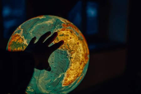 Explorando el mundo: Una mano llega a un globo brillante en una habitación oscura. Viaje educativo: Globo luminoso y busca nombres de países.Geografía interactiva.Búsqueda por país.