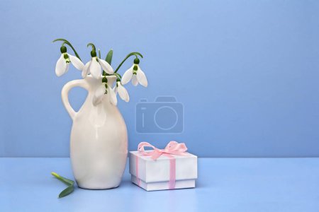 Strauß weiße Blumen Schneeglöckchen in Porzellanvase, Geschenkbox auf blauem Tisch auf blauem Hintergrund mit Platz für Text. Frühlingsdekoration