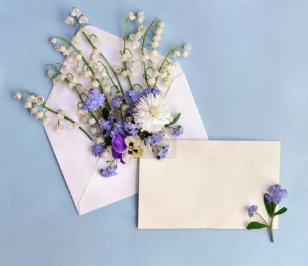 White flowers Lily of the valley ( Convallaria, May bells, may lily ), viola, forget-me-not, daisy, postal envelope with paper card note with space for text on a blue background. Top view, flat lay