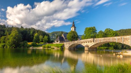 Téléchargez les photos : Longue exposition paysage naturel ensoleillé avec lac de montagne en Slovénie. beau lac Bohinj avec un ciel parfait en été. vue sur lac calme avec eau azur, église et pont en pierre sur fond. - en image libre de droit