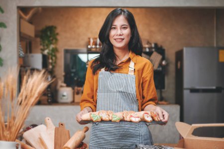 Téléchargez les photos : Femme asiatique aux cheveux longs portant une blouse jaune et un tablier debout tenant une plaque de pain de cuisson avec un visage souriant regardant la caméra à l'intérieur de la cuisine. - en image libre de droit