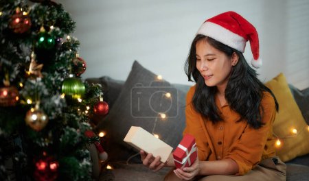 Téléchargez les photos : Belle femme dans une chemise jaune et une boîte cadeau d'ouverture de chapeau de Père Noël rouge avec une expression heureuse et excitée sur le cadeau de Noël ou du Nouvel An. - en image libre de droit