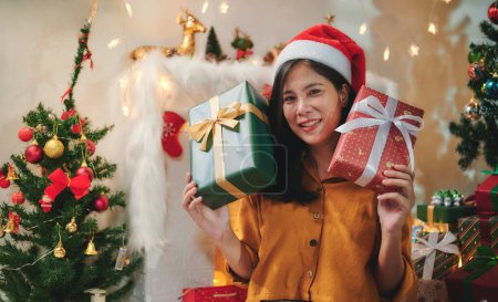 Téléchargez les photos : Belle femme aux cheveux longs portant une chemise jaune et un chapeau rouge Santa regarde la caméra avec un visage souriant heureux tout en tenant une grande boîte cadeau reçue à l'occasion de Noël dans les deux mains. - en image libre de droit