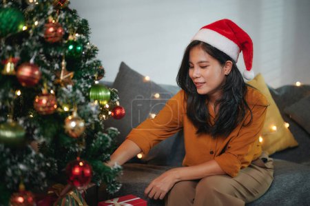 Téléchargez les photos : Belle femme portant un chapeau de Père Noël décoré l'arbre de Noël à l'intérieur de la maison avec des boules colorées, des boîtes-cadeaux et des ampoules colorées avant la veille de Noël pour célébrer la saison des fêtes. - en image libre de droit