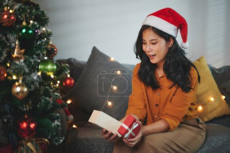 Téléchargez les photos : Belle femme dans une chemise jaune et une boîte cadeau d'ouverture de chapeau de Père Noël rouge avec une expression heureuse et excitée sur le cadeau de Noël ou du Nouvel An. - en image libre de droit