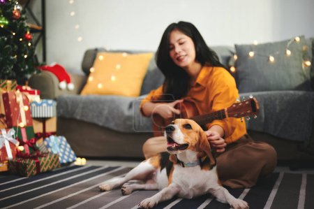 Téléchargez les photos : Bonne femme célébrant Noël ou le Nouvel An avec beagle chiot et guitare préférée à l'intérieur de la maison décorée avec arbre de Noël, boîte cadeau et ampoule. - en image libre de droit