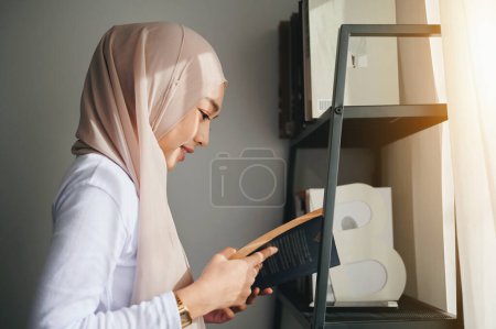 Téléchargez les photos : Une femme portant un foulard islamique en hijab lit un livre près d'une étagère près de la fenêtre. - en image libre de droit