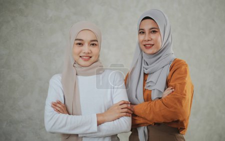 Foto de Dos mujeres asiáticas islámicas con vestidos formales y pañuelos para la cabeza hijab están de pie con los brazos cruzados sonriendo con confianza y amables mirando a la cámara. - Imagen libre de derechos