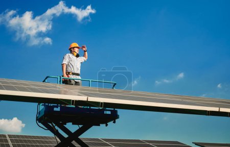 Téléchargez les photos : Homme ingénieur installateur de panneaux solaires debout sur le téléphérique portant un casque jaune et tenant exercice, impatient parmi de nombreux panneaux solaires par temps ensoleillé avec un ciel clair. Production d'énergie solaire. - en image libre de droit