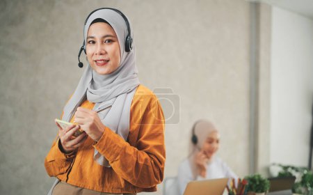 Téléchargez les photos : Femme musulmane portant un foulard en hijab et un casque debout à l'intérieur du bureau, concept de service après-vente, télévente et employés fournissent des informations en ligne. - en image libre de droit