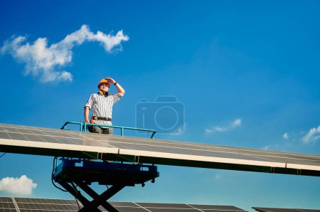 Téléchargez les photos : Homme ingénieur installateur de panneaux solaires debout sur le téléphérique portant un casque jaune et tenant exercice, impatient parmi de nombreux panneaux solaires par temps ensoleillé avec un ciel clair. Production d'énergie solaire. - en image libre de droit