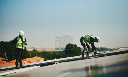 Téléchargez les photos : Deux ingénieurs en installation solaire hommes portant des combinaisons de protection travaillent sur le toit d'un immeuble de grande hauteur. - en image libre de droit