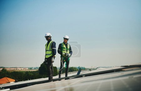 Foto de Dos ingenieros de instalaciones solares masculinos con trajes de protección se paran en el techo de un edificio de gran altura. - Imagen libre de derechos