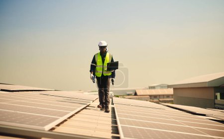 Téléchargez les photos : Homme ingénieur portant un équipement de protection en hauteur marchant tenant ordinateur portable sur le toit d'un bâtiment pour vérifier l'installation de panneaux solaires. - en image libre de droit