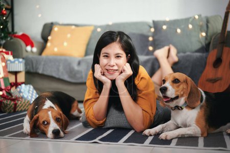 Téléchargez les photos : Long-haired Asian woman in a bright yellow blouse is lying on the floor and cheerfully with her two favorite beagle puppies in the living room. - en image libre de droit