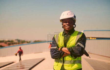 Téléchargez les photos : African engineer man installing solar panels wearing protective gear and a white helmet is standing arm crossed on the roof. - en image libre de droit