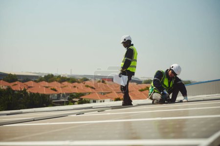 Téléchargez les photos : Deux hommes de nationalités différentes portant des vêtements de protection pour travailler sur des immeubles de grande hauteur installent des panneaux solaires sur le toit d'un bâtiment. - en image libre de droit