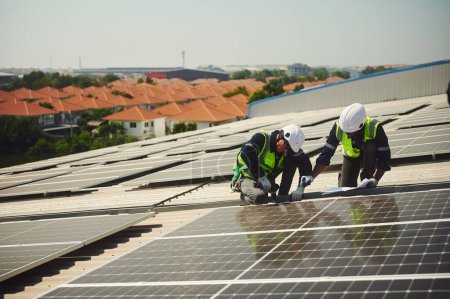 Téléchargez les photos : Deux ingénieurs de nationalités différentes portant des vêtements de protection pour travailler sur des immeubles de grande hauteur installent des panneaux solaires sur le toit d'un bâtiment. - en image libre de droit