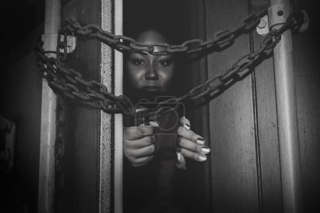 Téléchargez les photos : Photo noir et blanc femme afro-américaine sans défense piégée dans un conteneur illicite verrouillé avec une chaîne pitoyable et cadenas : Concept de traite des êtres humains - en image libre de droit