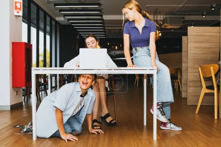 Photo for Female teacher and naughty student pupils : Young beautiful teacher sits desk without complaining in the school library with boys and girls having fun sit on the table and below them mischievously. - Royalty Free Image