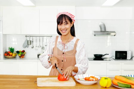 Téléchargez les photos : Chef novice : Portrait jeune femme asiatique de bonne humeur tranchant tomates et légumes frais avec sourire, cuisiner des aliments savoureux et nutritifs à la maison dans la cuisine moderne et équipement de cuisine propre. - en image libre de droit