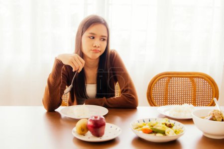 Foto de Joven mujer asiática hambrienta pero aburrida comiendo en la mesa llena de comida blanda sentada mirando la comida con aburrimiento y teniendo anorexia, no feliz de comer y no tener buen apetito. - Imagen libre de derechos